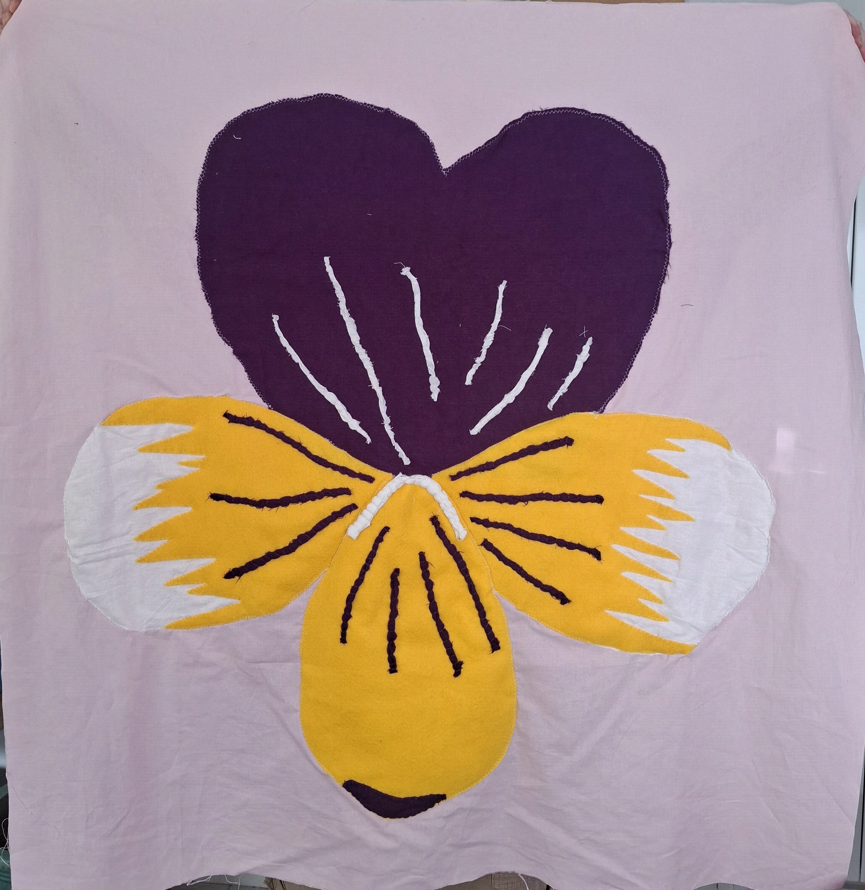 a purple, yellow and white viola triocolour made of fabric sewn onto a large pink square of fabric